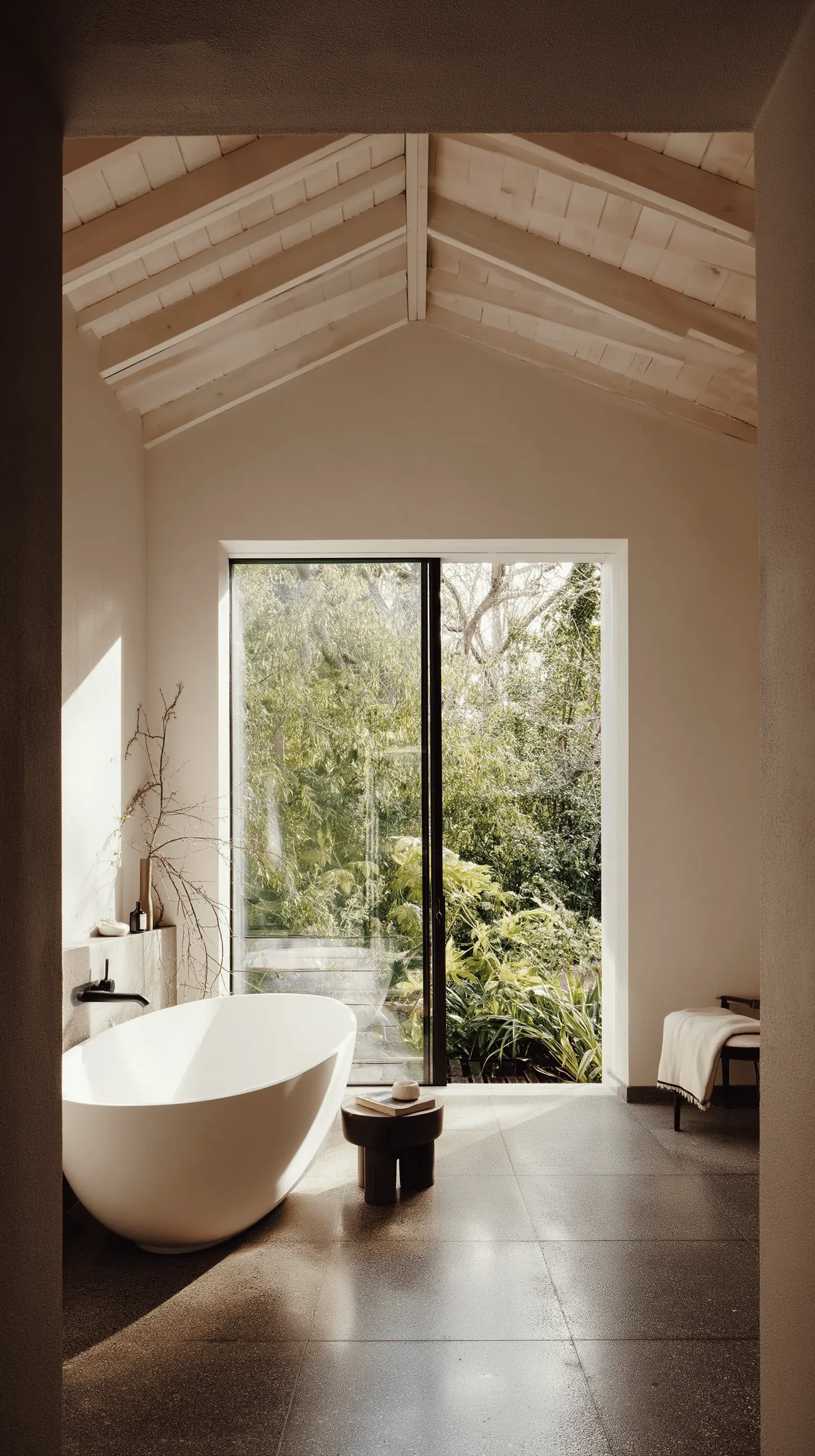 A narrow bathroom with a freestanding oval bath beneath a pitched white timber ceiling, a glazed door opening to greenery