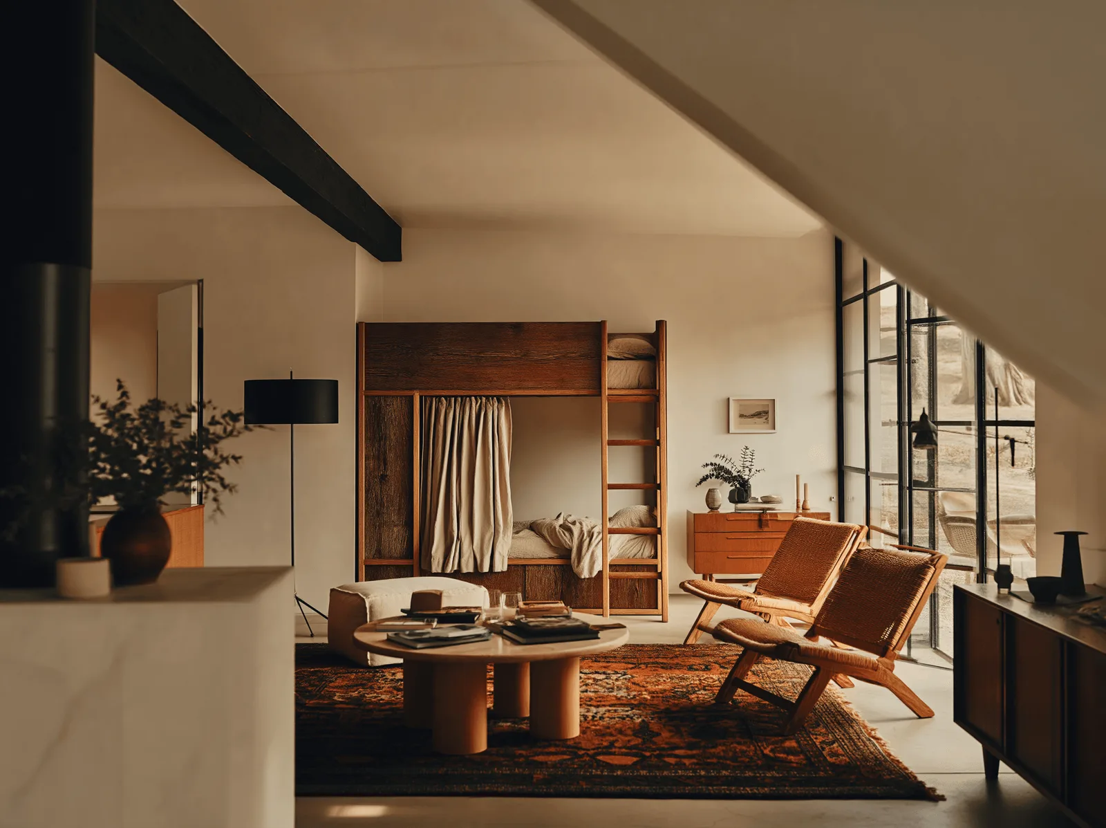An open-plan living room with exposed black steel beams, a stone kitchen bench, timber armchairs and a patterned rug